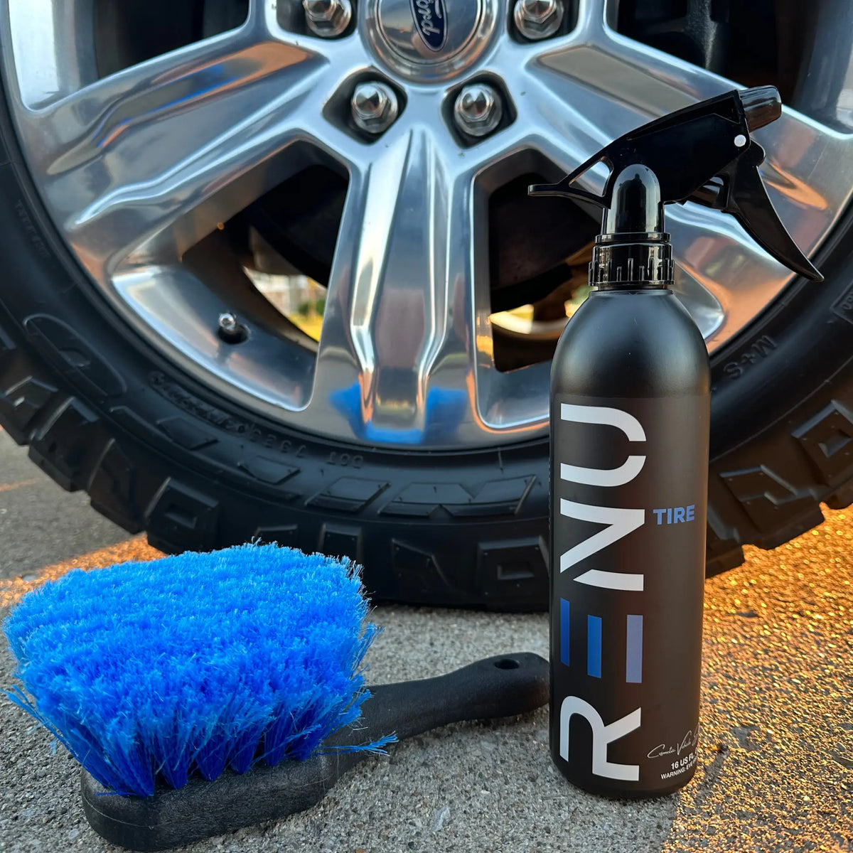 A black spray bottle labeled 'RENU TIRE' stands next to a bright blue tire brush on a paved surface, with a car wheel in the background.