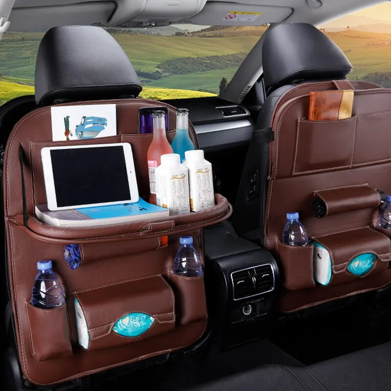 Brown faux-leather car rear seat organizer covers installed on both front seats inside a vehicle, shown holding a tablet, bottles, papers, and other items. The car interior and a scenic road are visible in the background.