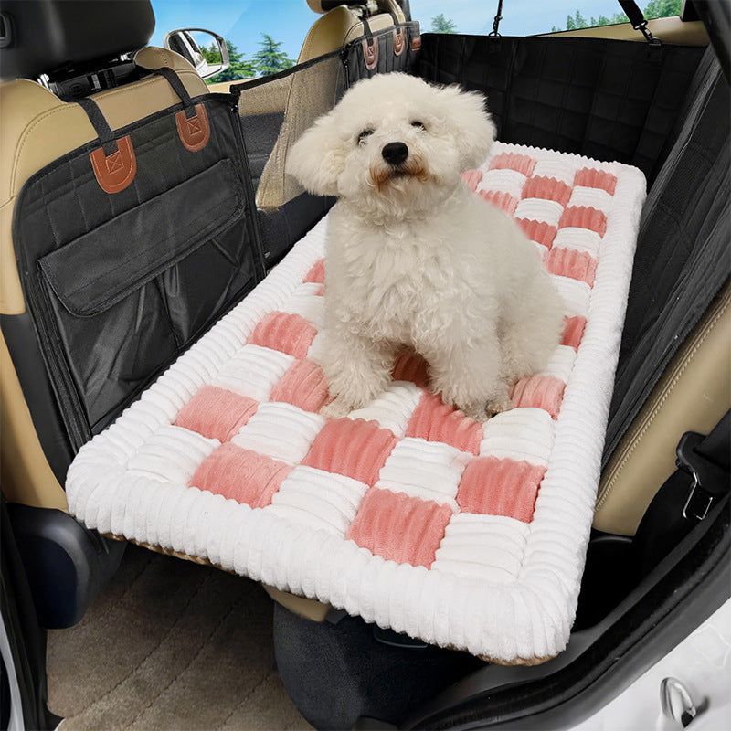 Small white dog sitting on a pink and cream checkered plush mat on a car back seat