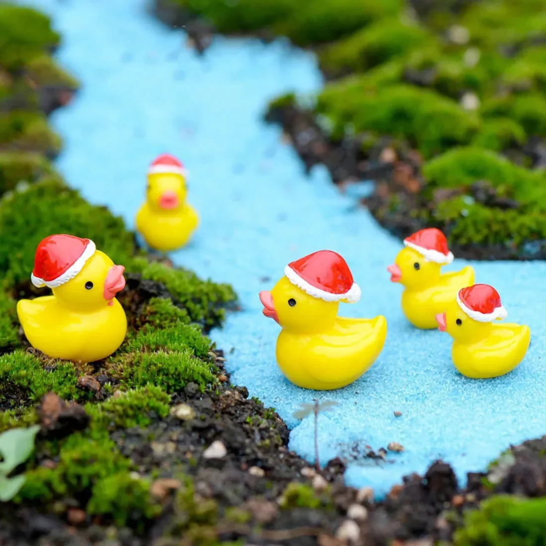 Four small yellow rubber ducks wearing red Santa hats are arranged on a blue path surrounded by green moss and dark soil.