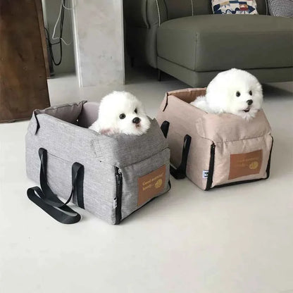 Two small white fluffy dogs sit inside pet booster car seats, one gray and one beige, placed on a light floor in a living room setting.