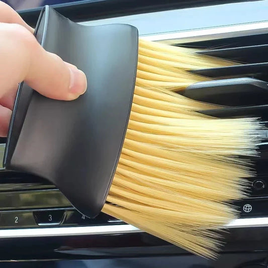 A hand holds a black car air vent cleaning brush with beige bristles against a car air vent.